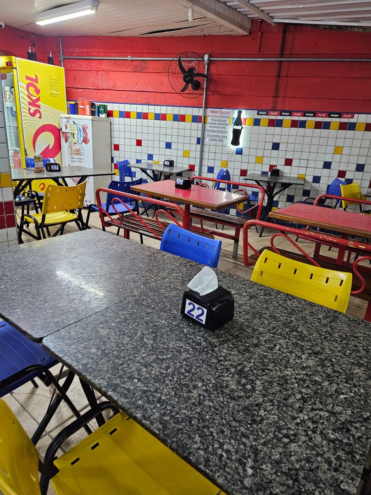 Salão com parede de azulejo branco, vermelho, amarelo e azul, mesas de granito e cadeiras coloridas